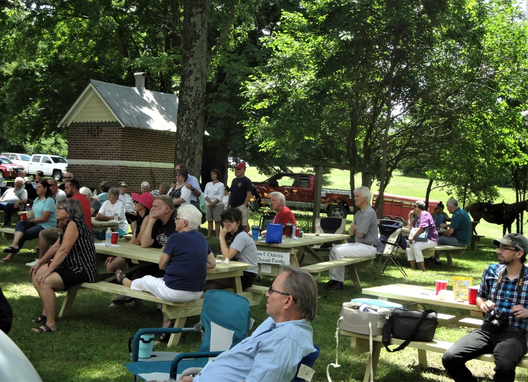 A crowd of people at picnic tables on the grounds of the CHC with the brick outhouse in the background